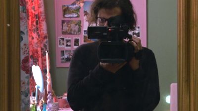 Film still from 'Field Reports' (2025) by Jon Beaton. The filmmaker is seen in the mirror standing in a pink bedroom holding a camcorder.